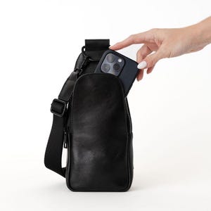 May include: Black leather sling bag with a zippered main compartment and adjustable strap. A hand is placing a black smartphone into the bag's pocket. The bag is designed to be worn across the body.