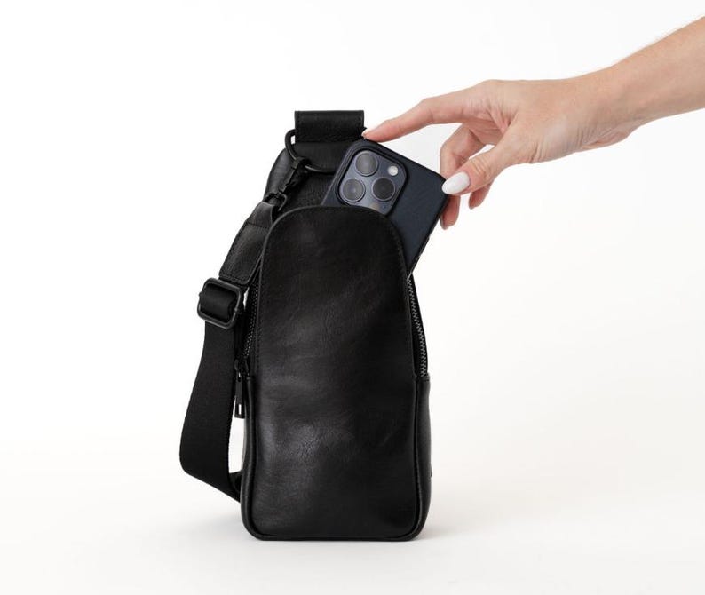 May include: Black leather sling bag with a zippered main compartment and adjustable strap. A hand is placing a black smartphone into the bag's pocket. The bag is designed to be worn across the body.