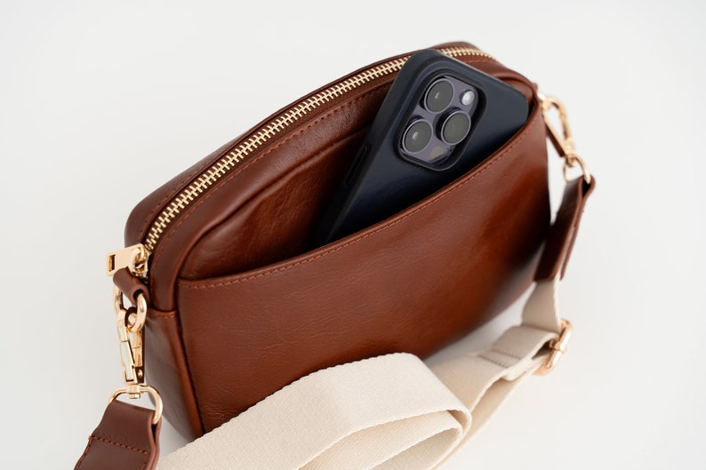 May include: A brown leather crossbody bag with a cream canvas strap and a gold zipper. A black smartphone is inside the bag with the camera lens visible.