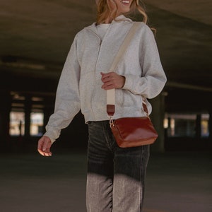 May include: A woman wearing a gray zip-up hoodie, a brown beanie, and black sunglasses is carrying a brown leather crossbody bag with a cream strap.