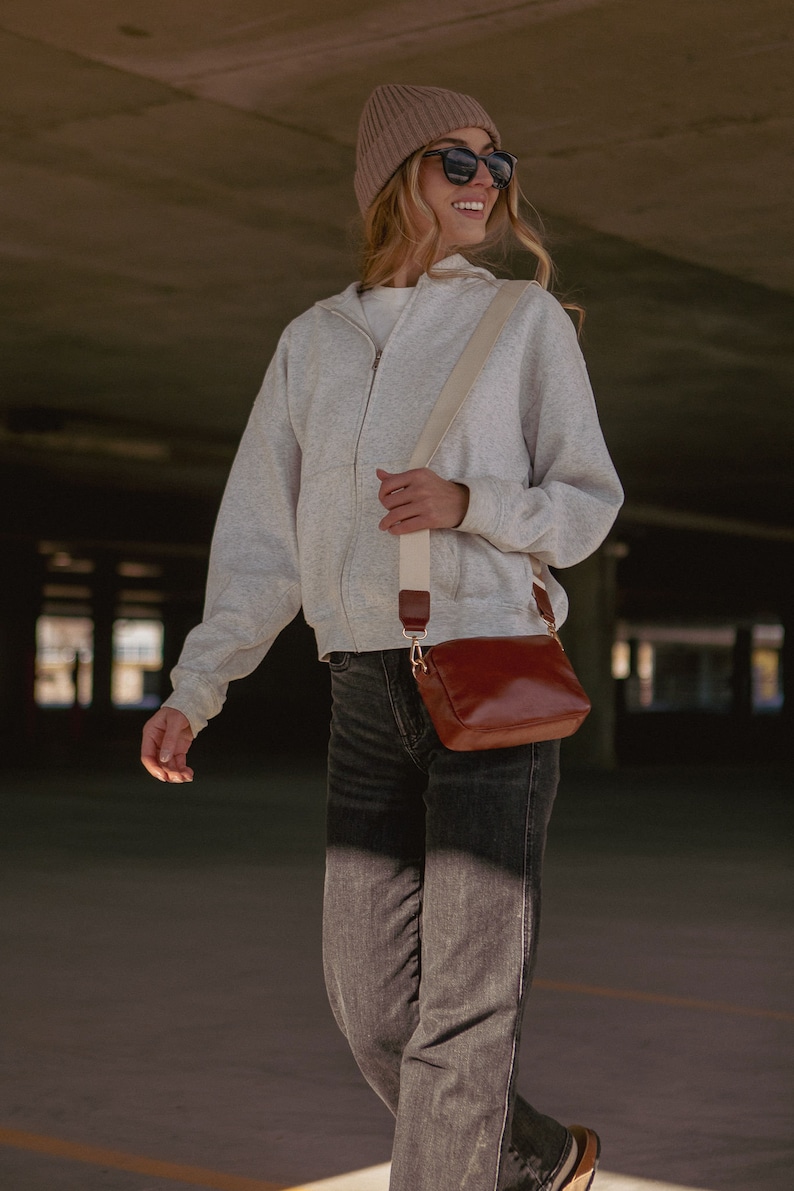May include: A woman wearing a gray zip-up hoodie, a brown beanie, and black sunglasses is carrying a brown leather crossbody bag with a cream strap.
