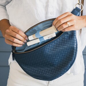 May include: A navy blue woven leather crossbody bag with a light blue interior. The bag is open, revealing two cosmetic tubes and a beige tube. The bag has a white strap and gold-colored hardware.