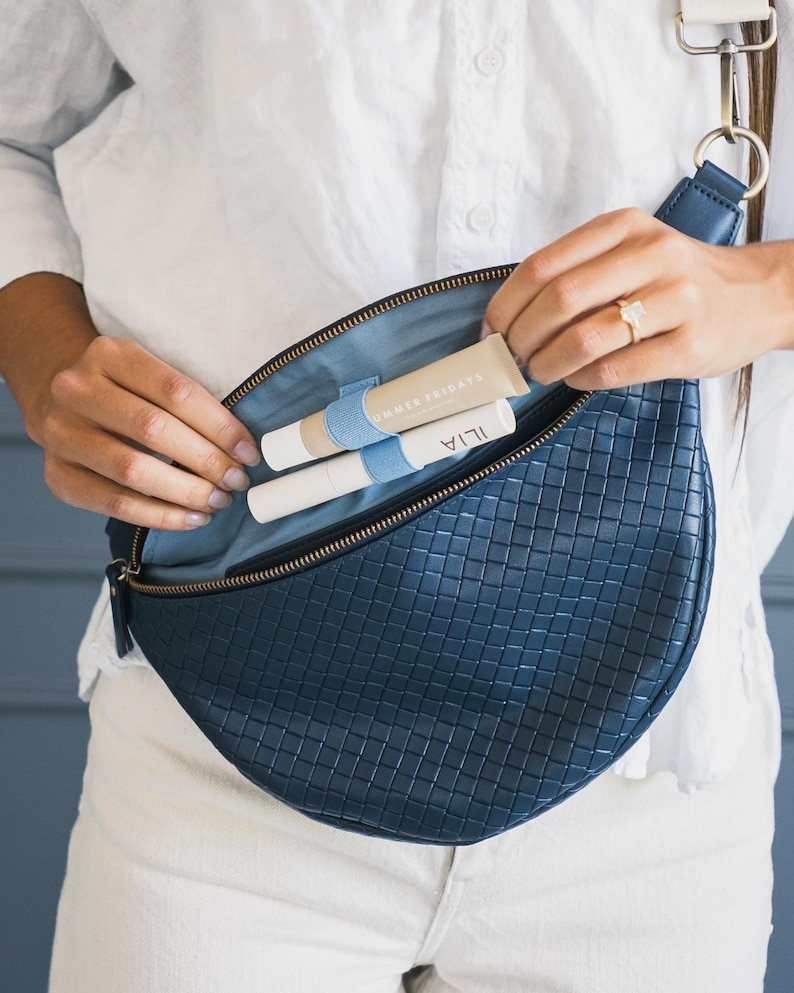 May include: A navy blue woven leather crossbody bag with a light blue interior. The bag is open, revealing two cosmetic tubes and a beige tube. The bag has a white strap and gold-colored hardware.