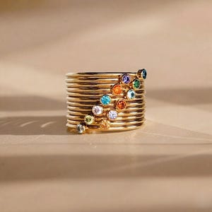 May include: A collection of gold-coloured rings, each with a thin band. Each ring is decorated with a small, colourful gemstone. The gemstones are in various colours, including blue, green, orange, and purple. The rings are stacked together, creating a layered effect.