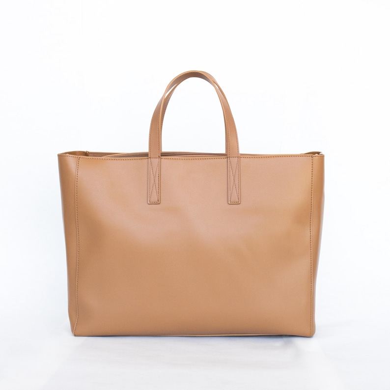 May include: A tan leather tote bag with two top handles and a rectangular shape. The bag has a simple, minimalist design and is suitable for everyday use. The bag is made of a smooth, solid material.