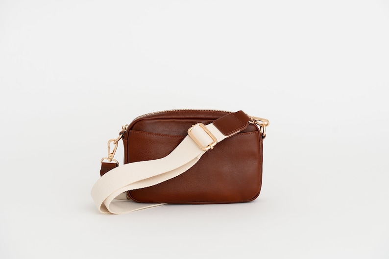 May include: A brown leather crossbody bag with a white adjustable strap and gold hardware.