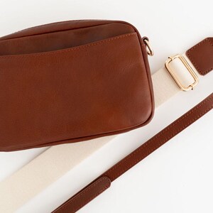 May include: A brown leather crossbody bag with a cream and brown adjustable strap. The bag has a flap closure and a small pocket on the front.