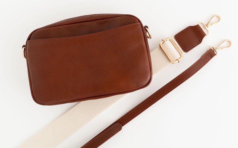 May include: A brown leather crossbody bag with a cream and brown adjustable strap. The bag has a flap closure and a small pocket on the front.