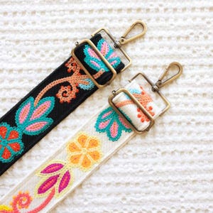 May include: Two embroidered bag straps with metal hardware. One strap is black with teal, pink, and orange floral designs. The other is off-white with similar colorful floral embroidery. Both straps have gold-colored metal clasps.