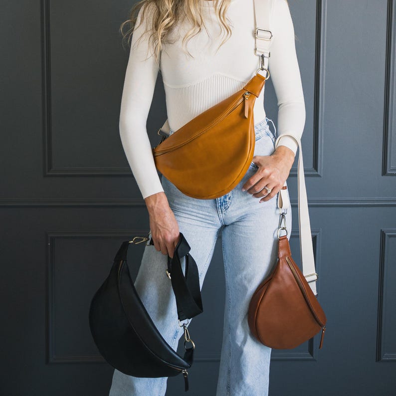 May include: Three leather crossbody bags in different colors: tan, black, and brown. The bags have a curved shape with a zipper closure and adjustable straps. The tan bag is worn across the body, while the black bag is held in the hand, and the brown bag hangs from the waist.