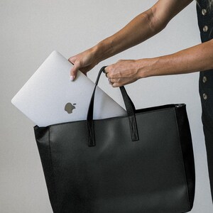 May include: A black tote bag with a rectangular shape and two handles. A silver laptop with the Apple logo is being placed inside the bag. The bag has a simple, modern design, suitable for carrying everyday essentials.