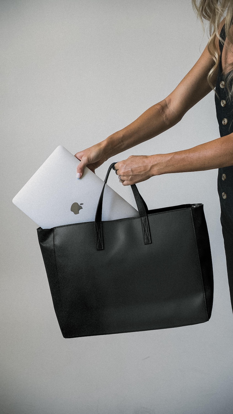 May include: A black tote bag with a rectangular shape and two handles. A silver laptop with the Apple logo is being placed inside the bag. The bag has a simple, modern design, suitable for carrying everyday essentials.
