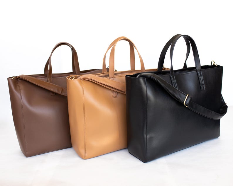 May include: Three leather tote bags in shades of brown and black. The bags have top handles and a rectangular shape. The black bag has a shoulder strap. The bags are displayed against a white background.