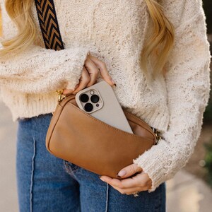 May include: A brown leather crossbody bag with a gold chain strap. A white phone with a black camera lens is in the bag.