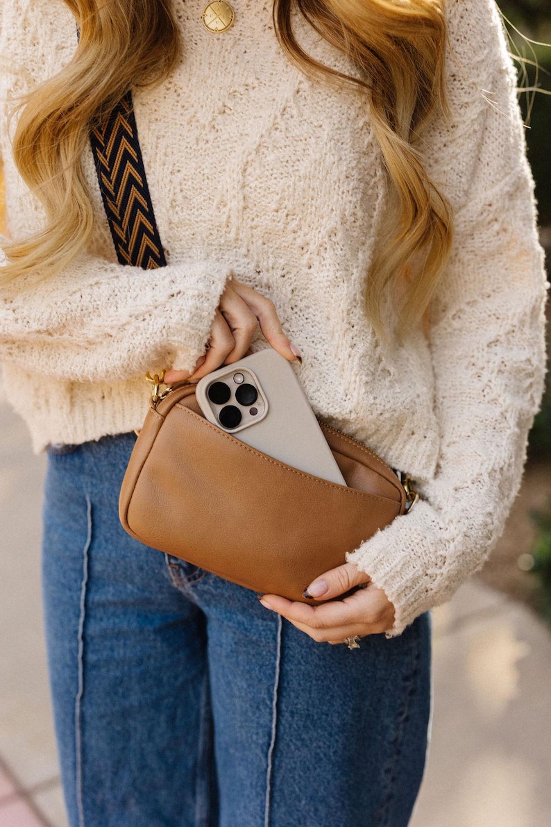 May include: A brown leather crossbody bag with a gold chain strap. A white phone with a black camera lens is in the bag.