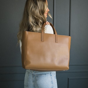 May include: A large, rectangular brown tote bag with two top handles. The bag is made of a smooth material and has a simple, classic design. The bag is being held by a person wearing a white top and blue jeans.