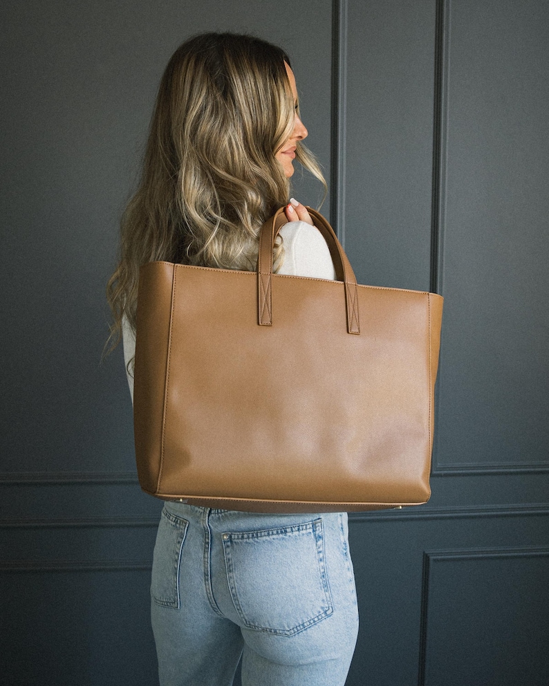 May include: A large, rectangular brown tote bag with two top handles. The bag is made of a smooth material and has a simple, classic design. The bag is being held by a person wearing a white top and blue jeans.