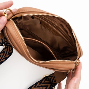 May include: A tan leather crossbody bag with a gold zipper and a black and brown patterned strap.