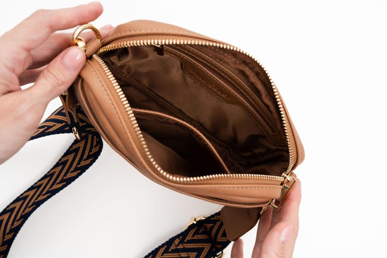 May include: A tan leather crossbody bag with a gold zipper and a black and brown patterned strap.