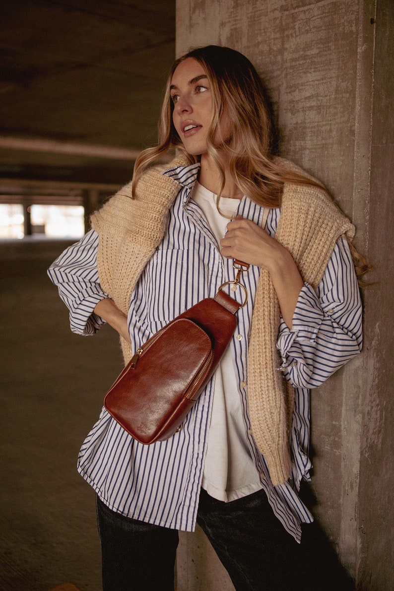 May include: A brown leather crossbody bag with a gold zipper and hardware. The bag is worn over a white t-shirt and a blue and white striped button-down shirt. A beige sweater is draped over the shoulders.