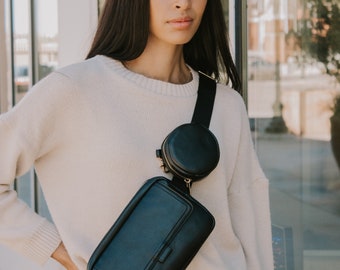 May include: A black leather fanny pack with a round, black pouch attached. The fanny pack has a gold zipper and buckle. The model is wearing a white sweater and beige pants.