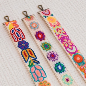 May include: Three colorful embroidered straps with floral and geometric designs. The straps are cream-colored with vibrant embroidery in shades of yellow, orange, pink, blue, and green. Each strap has a metal clasp at the top.