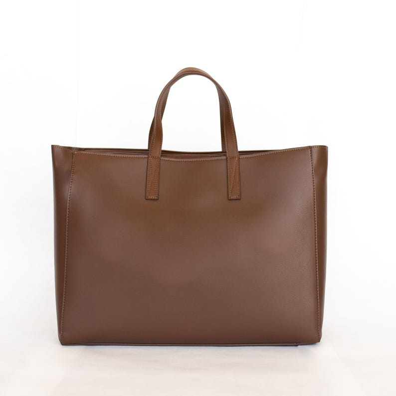 May include: A brown leather tote bag with two handles and a rectangular shape. The bag has a smooth texture and a simple design, suitable for carrying various items. The handles are attached to the top of the bag.