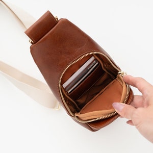 May include: A brown leather sling bag with a cream-colored strap. The bag has a zippered main compartment and a smaller zippered pocket. The bag is open, revealing the interior and contents.