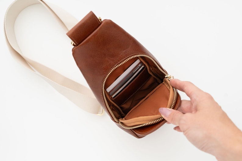 May include: A brown leather sling bag with a cream-colored strap. The bag has a zippered main compartment and a smaller zippered pocket. The bag is open, revealing the interior and contents.