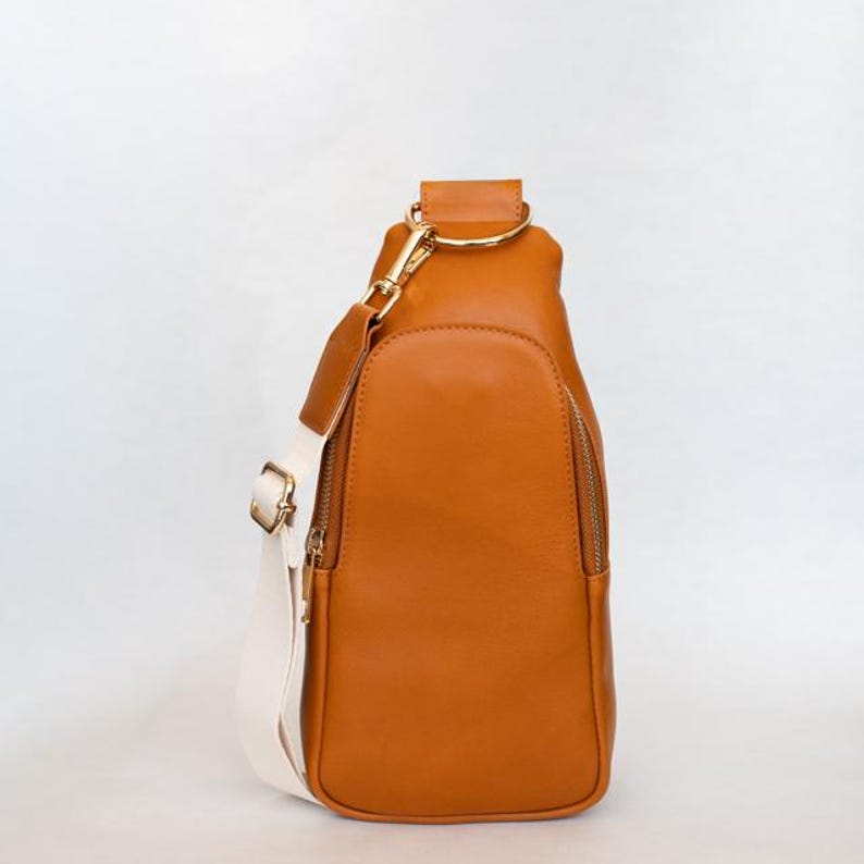 May include: A brown leather sling bag with a white strap and gold-colored hardware. The bag has a zippered pocket on the front and a gold-colored ring at the top for attaching the strap. The bag is approximately 12 inches tall.