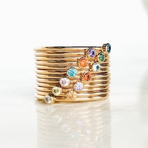 May include: A stack of gold rings with colorful gemstones. The rings are arranged in a rainbow pattern, with each ring featuring a different colored gemstone.