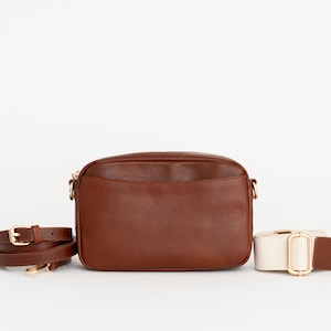 May include: A brown leather crossbody bag with a gold zipper and a matching brown strap. A second, off-white strap with a gold buckle is also included.