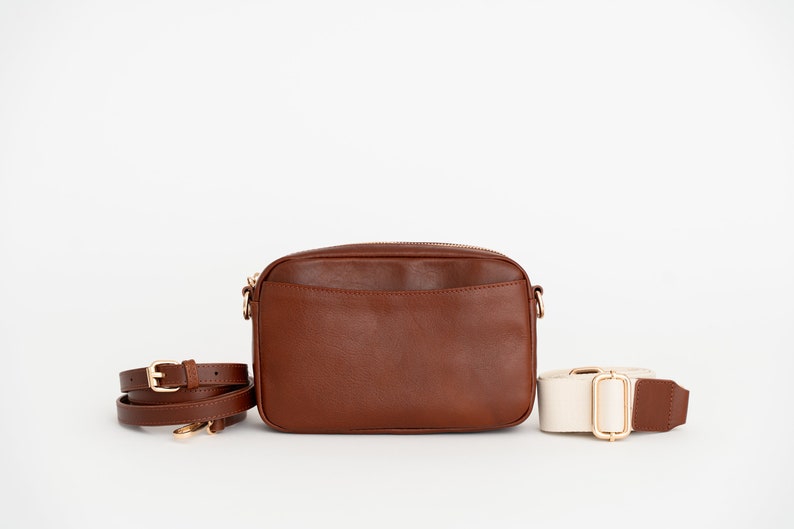 May include: A brown leather crossbody bag with a gold zipper and a matching brown strap. A second, off-white strap with a gold buckle is also included.