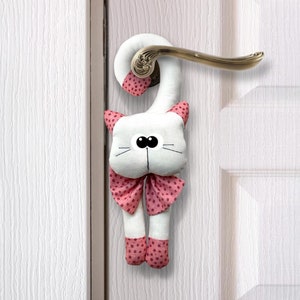 Puede incluir: Tapón de puerta de felpa blanca con forma de gato con un lazo rosa de lunares colgando de una puerta blanca.