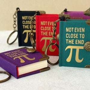 May include: A collection of colorful mini-book keychains. Each keychain features a book with the text "NOT EVEN CLOSE TO THE END" and the Pi symbol. The books come in various colors, including purple, teal, red, and black.