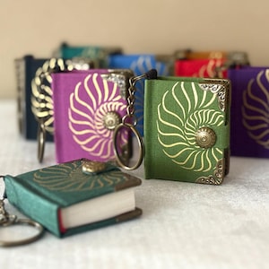 May include: A collection of small, colourful book keyrings. The miniature books are in various colours, such as teal, purple, and green, with a gold spiral design on the cover. Each book has a metal clasp and a keyring.