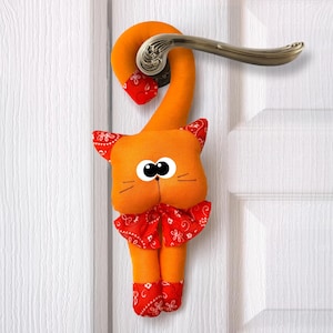 Może przedstawiać: Pomarańczowy, materiałowy, kot-stopka do drzwi z czerwoną chustką, wiszący na białej klamce.