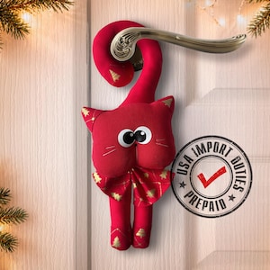 May include: A red fabric cat door hanger with a long curved tail, large white eyes, and a bow tie featuring a gold Christmas tree pattern. The cat has matching gold tree prints on its ears and feet, and it hangs from a metallic door handle.