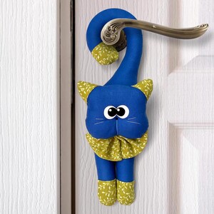 Może przedstawiać: Stoper do drzwi w kształcie niebieskiego kota z żółtymi i zielonymi akcentami. Kot ma duże czarne oczy i biały nos. Kot wisi na białych drzwiach.