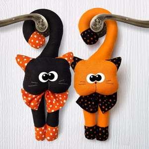 May include: Two cat-shaped door hangers in black and orange. The black cat has orange polka dots, the orange cat has black polka dots. Both have large eyes, whiskers, and bow ties. They hang from a bronze door handle.