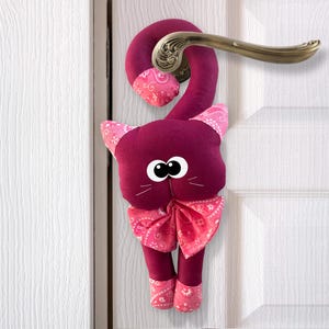 Può includere: Un fermaporta di peluche color borgogna a forma di gatto con un fiocco e una coda in tessuto a fantasia rosa e bianca. Il gatto ha grandi occhi neri e un naso bianco.