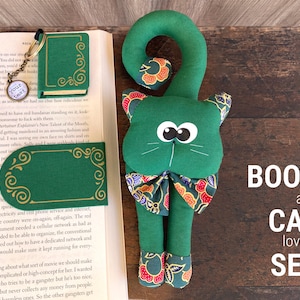 Może przedstawiać: Zielony zakładek w kształcie kota z kwiatową kokardką i zielona książka ze złotym zakładek. Kot i książka znajdują się na drewnianej powierzchni z tekstem "BOOK and CAT lovers SET".