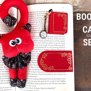 Könnte beinhalten: Ein rotes Buch- und Katzen-Lesezeichen-Set mit goldenen Akzenten. Das Buch ist eine Miniaturversion mit einem goldgeprägten Design. Die Katze ist ein Plüschtier mit einem roten Körper und einem schwarz-weißen Bandana. Das Set befindet sich auf einem Buch mit dem Text "BOOK and CAT lovers SET".