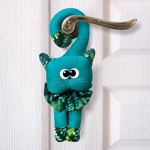 Peut inclure: Un arrêt de porte en forme de chat bleu sarcelle avec un motif floral vert et blanc. Le chat a de grands yeux noirs et un nez noir. Le chat est accroché à une porte blanche.