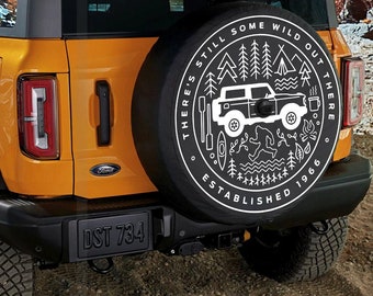 May include: A yellow Ford Bronco with a black spare tire cover. The spare tire cover has a white graphic design with the text "There's Still Some Wild Out There Established 1966".