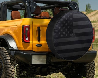 May include: A yellow Ford Bronco with a black spare tire cover featuring an American flag design. The vehicle is parked on a grassy hill.
