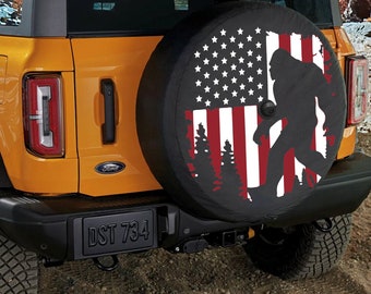 May include: A yellow Ford Bronco with a spare tire cover featuring an American flag design with a silhouette of Bigfoot walking in front of a forest. The spare tire cover is black with red and white accents. The license plate reads "DST 734".
