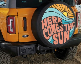 May include: A yellow Ford Bronco with a spare tire cover that says "Here Comes The Sun" in a retro font with a sun and wave design.