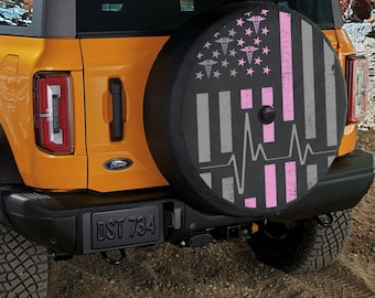 May include: A black spare tire cover with a distressed American flag design. The flag has a pink stripe down the right side and a pink heartbeat graphic. The flag also has medical caduceus symbols in place of the stars.