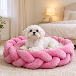May include: A small white dog with long fur resting on a round, bright pink pet bed. The bed features a thick, plush, braided border that forms a circular cushion, providing a soft and textured surface for the animal to lie upon.